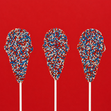 Three solid milk chocolate ice cream cone pops covered entirely in red, white and blue nonpareil beads on white sticks against a red background, patriotic Fourth of July treat by Nibbles and Bits Scranton PA.