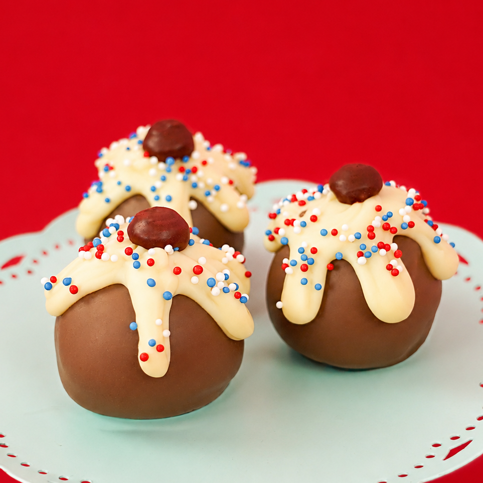 Three patriotic sundae milk chocolate ganache truffles on a mint green cake stand with red background, each topped with dripping white chocolate, red, white and blue nonpareils and a dark chocolate cherry, handmade Fourth of July gift by Nibbles and Bits Scranton PA.