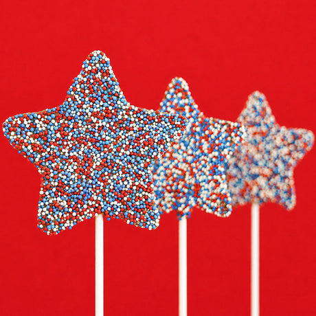 Three solid milk chocolate star lollipops covered entirely in red, white and blue nonpareil candy beads on white sticks against a bold red background, patriotic Fourth of July treat by Nibbles and Bits Scranton PA.