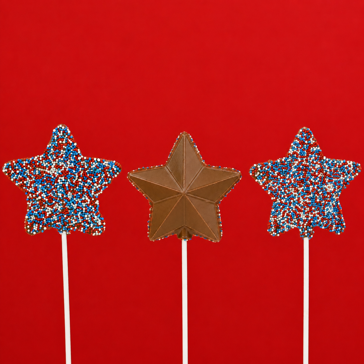 Three solid milk chocolate patriotic star pops on white sticks against a bold red background showing both the red, white and blue nonpareil covered side and the detailed geometric faceted molded star side, handmade Fourth of July treat by Nibbles and Bits Scranton PA.