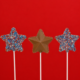 Three solid milk chocolate patriotic star pops on white sticks against a bold red background showing both the red, white and blue nonpareil covered side and the detailed geometric faceted molded star side, handmade Fourth of July treat by Nibbles and Bits Scranton PA.