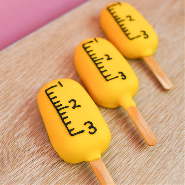 Peanut butter crème pop dipped in yellow chocolate and hand-detailed to look like a school ruler with black measurement markings and number increments on a stick, handmade back to school gift by Nibbles and Bits Scranton PA.