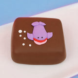 Milk chocolate s'more decorated with an adorable purple fondant whale with red smile and white bubble details on a white plate with blue background, handmade ocean themed treat by Nibbles and Bits Scranton PA.