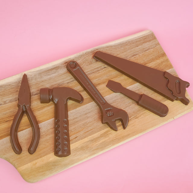 Five small solid milk chocolate tools including pliers hammer wrench screwdriver and hand saw displayed on a rustic wood board with pink background, handmade novelty Father's Day gift by Nibbles and Bits Scranton PA.