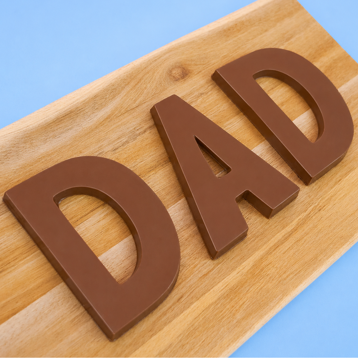Three large solid milk chocolate letters spelling DAD displayed on a rustic wood board with blue background, handmade Father's Day gift by Nibbles and Bits Scranton PA.