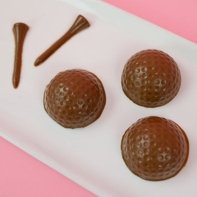 Three solid milk chocolate golf balls with realistic dimple texture and three milk chocolate golf tees on a white rectangular plate with pink background, handmade golf gift set by Nibbles and Bits Scranton PA.