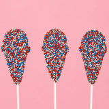 Three solid milk chocolate ice cream cone pops covered entirely in red, white and blue nonpareil beads on white sticks against a pink background, patriotic Fourth of July treat by Nibbles and Bits Scranton PA.
