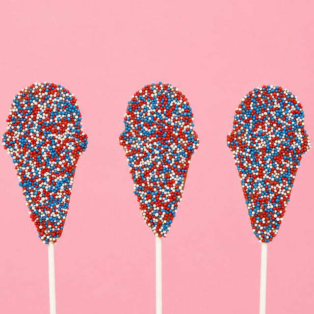 Three solid milk chocolate ice cream cone pops covered entirely in red, white and blue nonpareil beads on white sticks against a pink background, patriotic Fourth of July treat by Nibbles and Bits Scranton PA.