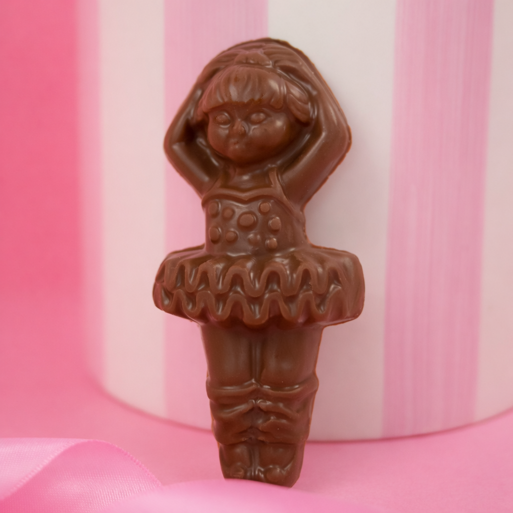 Milk chocolate ballerina shaped candy standing upright in front of a pink and white striped background, with a soft pink ribbon in the foreground.