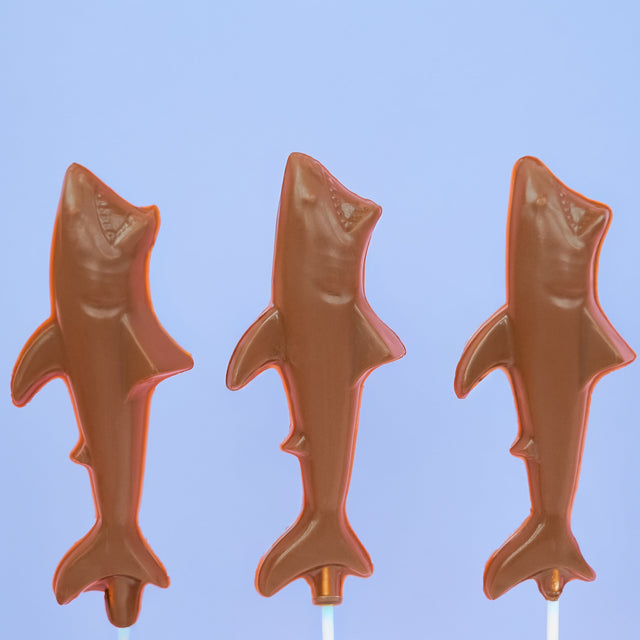 Three solid milk chocolate shark lollipops with detailed fin jaw and body molding on white sticks against a blue background, handmade ocean themed party favor by Nibbles and Bits Scranton PA.