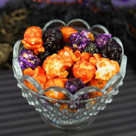 Glass pedestal bowl filled with orange, purple, and black glazed Halloween popcorn against a purple background.