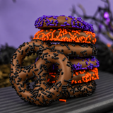 Seven milk chocolate covered pretzels with orange, purple and black sprinkles, lined up on a striped plate with a Halloween background. 