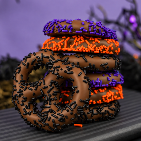 Seven milk chocolate covered pretzels with orange, purple and black sprinkles, lined up on a striped plate with a Halloween background. 