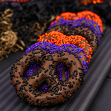 Seven milk chocolate covered pretzels with orange, purple and black sprinkles, lined up on a striped plate with a Halloween background. 
