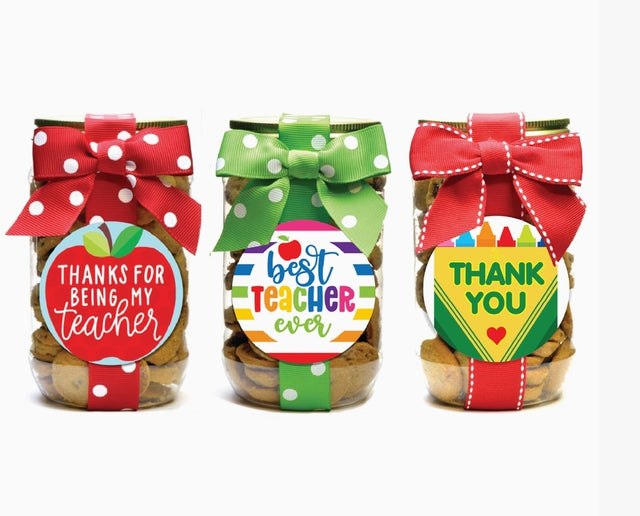 Three teacher appreciation mini chocolate chip cookie pint jars with decorative ribbons and labels reading Thanks for Being My Teacher, Best Teacher Ever, and Thank You with crayons, handmade gift by Nibbles & Bits.