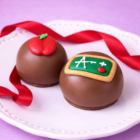 Two milk chocolate ganache truffles on a white plate with red satin ribbon — one topped with a red fondant apple, one with a green chalkboard A+ decoration, teacher appreciation gift by Nibbles & Bits.
