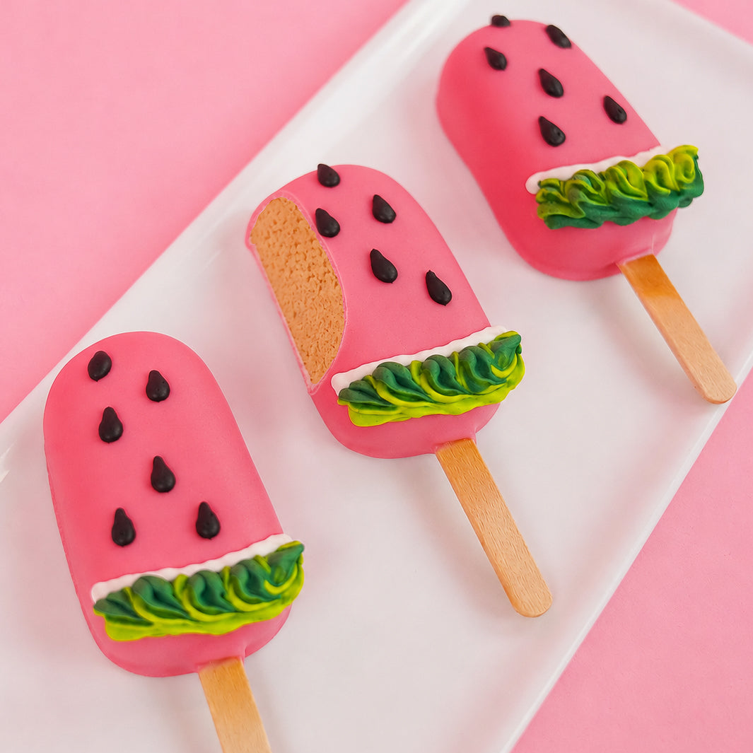 A white chocolate watermelon peanut butter pop revealing a creamy handmade peanut butter ganache center alongside two whole decorated pops on a white plate with pink background, gourmet summer treat by Nibbles and Bits Scranton PA.