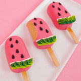 A white chocolate watermelon peanut butter pop revealing a creamy handmade peanut butter ganache center alongside two whole decorated pops on a white plate with pink background, gourmet summer treat by Nibbles and Bits Scranton PA.