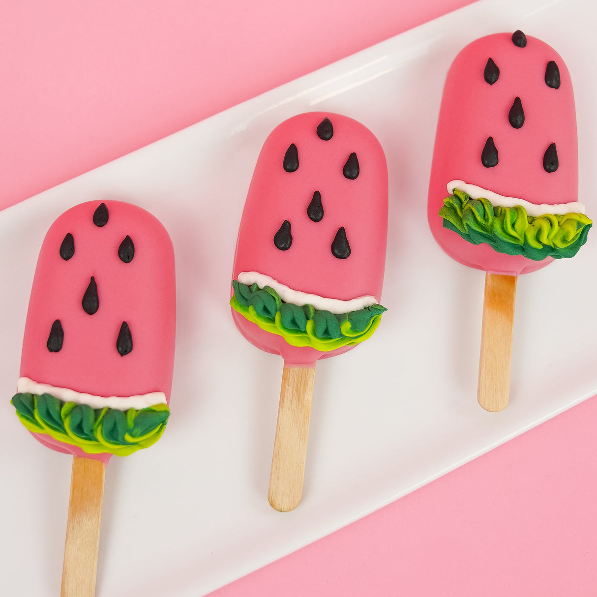 Three white chocolate watermelon peanut butter pops on a white rectangular plate with pink background, each coated in vivid pink white chocolate with black royal icing watermelon seeds and green and white ruffled rind detail, handmade summer treat by Nibbles and Bits Scranton PA.