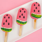 Three white chocolate watermelon peanut butter pops on a white rectangular plate with pink background, each coated in vivid pink white chocolate with black royal icing watermelon seeds and green and white ruffled rind detail, handmade summer treat by Nibbles and Bits Scranton PA.