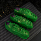 Three green chocolate Witch Fingers with textured knuckle creases and hand-painted purple almond nails on a striped surface.