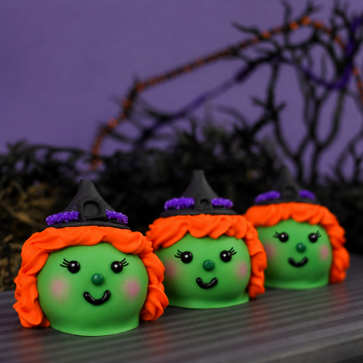 Three Witch Ganache Truffles with green chocolate faces, orange icing hair, blushing cheeks, and chocolate kiss witch hats on a striped surface.