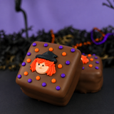 Two chocolate-covered s'mores decorated with orange and purple icing dots and a small witch face topper, photographed on a dark surface with a purple and black Halloween-themed background.