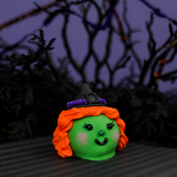 Witch Ganache Truffle with green chocolate faces, orange icing hair, blushing cheeks, and chocolate kiss witch hats on a striped surface.